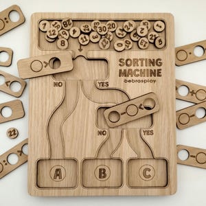 Könnte beinhalten: Holzsortiermaschinen-Puzzle mit dem Text "SORTING MACHINE" und "bebrasplay". Das Puzzle hat Schlitze für nummerierte Holzscheiben und Sortierpfade mit den Bezeichnungen "YES" und "NO". Enthält Holzsortierteile.