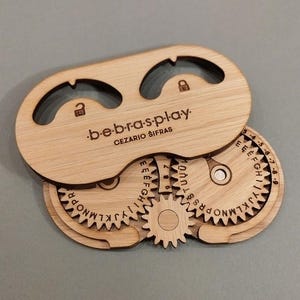 Può includere: Un puzzle in legno con un coperchio scorrevole e ingranaggi rotanti. Gli ingranaggi sono etichettati con lettere e numeri. Il coperchio ha il testo "bebras-play" e "CEZARIO ŠIFRAS".