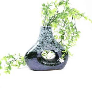 Può includere: Un vaso in ceramica blu scuro e verde con un'apertura circolare. Il vaso ha un motivo maculato sulla parte superiore ed è riempito con steli verdi e frondosi. Il vaso è un oggetto decorativo.