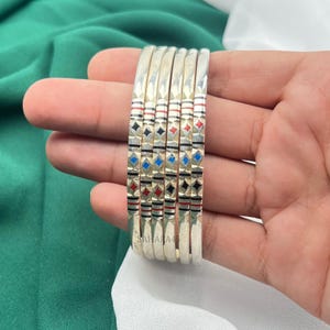 May include: A set of silver bangles with a geometric pattern of black, red, and blue diamonds. The bangles are stacked together, showcasing their design. The word "SAHARA45" is printed on the bangles.