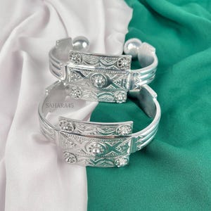 May include: Two silver-toned cuff bracelets with intricate floral and geometric designs. Each bracelet features a rectangular centerpiece with raised detailing and small, round embellishments. The bracelets are displayed on a white and green fabric background.