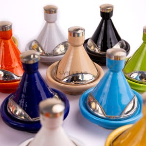 Może przedstawiać: Kolekcja ceramicznych tadżinów w różnych kolorach, w tym pomarańczowym, białym, czarnym, beżowym, niebieskim, jasnoniebieskim, zielonym i żółtym. Każda stożkowa pokrywka ma srebrny metalowy pasek i uchwyt. Na beżowym tadżinie widnieje napis SAHARA.