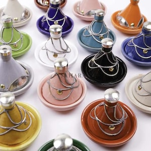 May include: A collection of colorful ceramic tagines, each with a conical lid and a circular base. The tagines come in various colors, including blue, green, orange, pink, and white, and are adorned with silver-colored metal accents.
