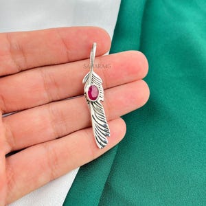May include: A silver-colored feather pendant with a vibrant red gemstone at its center. The pendant has detailed feather etching and a small loop at the top for hanging. The text "SAHARA45" is visible.