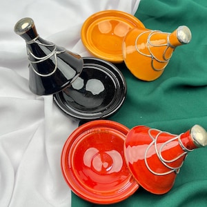 Op de afbeelding: Een verzameling kleurrijke tajine-potten in zwart, oranje en rood. De conische deksels zijn versierd met zilverkleurig metaal. De tajines worden gepresenteerd op een groen, wit en groen doek. Deze potten worden gebruikt om te koken.