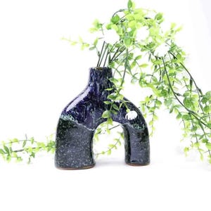 May include: A dark blue ceramic vase with a unique arch design. The vase has a speckled texture with green accents and a brown base. Green leafy stems with white flowers are arranged inside the vase, creating a decorative display.