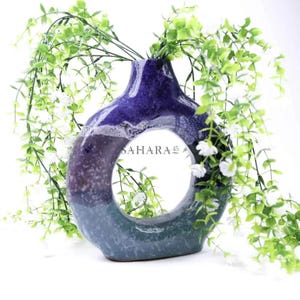 May include: A ceramic vase with a unique open circle design. The vase features a gradient of colors, transitioning from a deep blue at the top to shades of purple and teal at the base. The text "SAHARA 45" is visible on the side. Green and white floral accents are arranged around the vase.