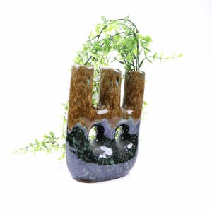 May include: A unique ceramic vase with three vertical openings, featuring a blend of brown, green, and blue hues. The vase has a textured surface and is filled with green leafy stems, adding a touch of nature to the design.