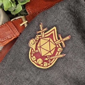 May include: A burgundy and gold embroidered patch featuring a geometric die, swords, and a key. The patch is sewn onto a dark gray fabric, with a brown leather belt and buckle in the background. The patch is likely for a role-playing game.