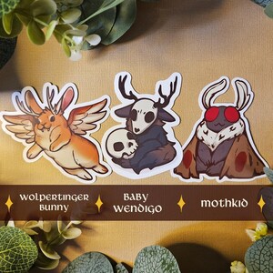 May include: Three fantasy creature sticker designs. The stickers depict a winged rabbit, a creature with antlers and a skull, and a moth-like creature. The stickers are labelled "wolpertinger bunny", "baby wendigo", and "mothkid".
