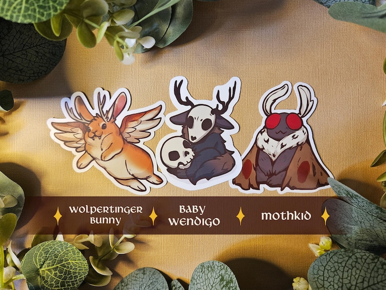 May include: Three fantasy creature sticker designs. The stickers depict a winged rabbit, a creature with antlers and a skull, and a moth-like creature. The stickers are labelled "wolpertinger bunny", "baby wendigo", and "mothkid".