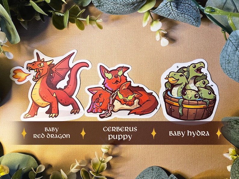 May include: Three fantasy creature stickers: a red dragon breathing fire, a Cerberus puppy, and a baby hydra in a wooden tub. Text on a banner reads "Baby Red Dragon", "Cerberus puppy", and "Baby hydra".