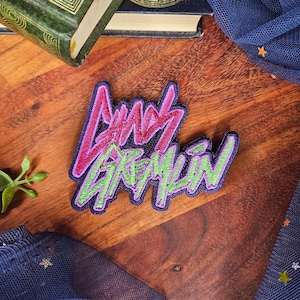 May include: Embroidered patch with the text "Lady Gremlin" in pink and green, outlined in purple. The patch is on a dark blue background. The patch is on a wooden surface.