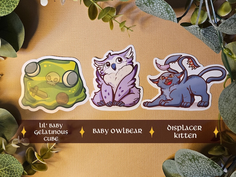 May include: Three fantasy creature stickers: a green gelatinous cube with a skull, a purple owlbear, and a blue displacer kitten. Text on the image reads "Lil' baby gelatinous cube", "baby owlbear", and "displacer kitten".