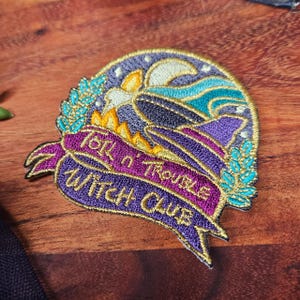 May include: Embroidered patch with a witch hat design, featuring the text "Talk n' Trouble Witch Club" on a purple banner. The patch has a gold border and includes a crescent moon, waves, and stars.