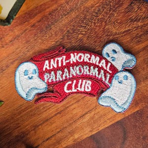 May include: Embroidered patch featuring the text "ANTI-NORMAL PARANORMAL CLUB" on a red banner, with three smiling ghost figures in white and blue. Ideal for adding to clothing or accessories.