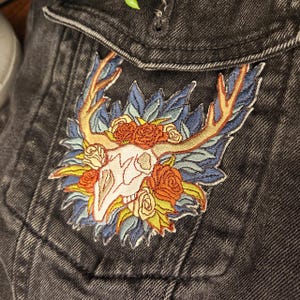 Peut inclure: Écusson brodé représentant un crâne blanc avec des bois, entouré de roses oranges, de feuilles bleues et de détails jaunes. Le patch est cousu sur une poche de veste en jean noire, avec un motif floral et nature détaillé.