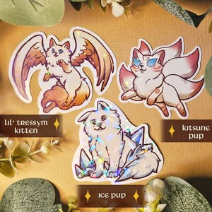May include: Three fantasy creature stickers: a winged cat labelled "Lil' tressym kitten", a fox-like creature labelled "kitsune pup", and a dog-like creature labelled "ice pup". The stickers are white with colourful accents and holographic details, set against a neutral background.