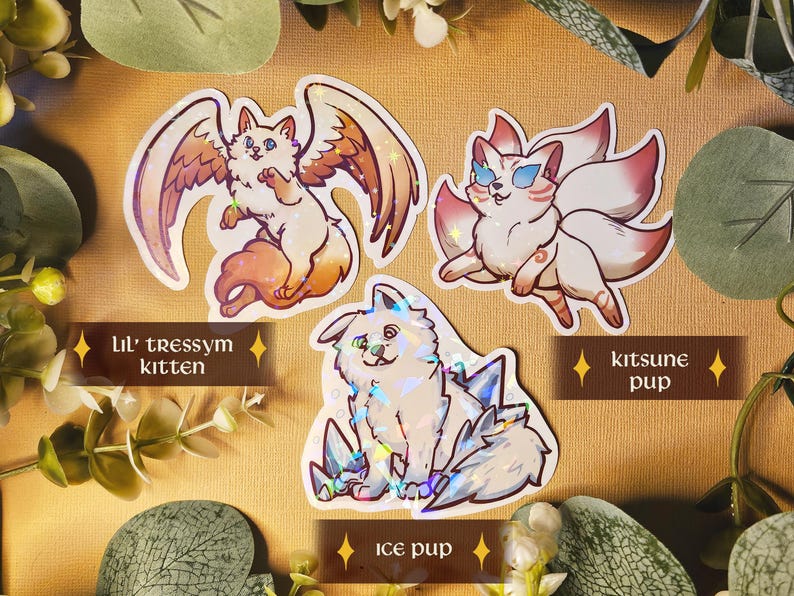 May include: Three fantasy creature stickers: a winged cat labelled "Lil' tressym kitten", a fox-like creature labelled "kitsune pup", and a dog-like creature labelled "ice pup". The stickers are white with colourful accents and holographic details, set against a neutral background.