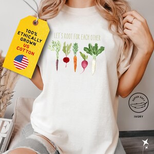 A white t-shirt with the text 'Let's root for each other' printed on it. The text is in green and brown. There are five watercolor illustrations of vegetables below the text: a carrot, a beet, a radish, a turnip, and a green onion. The t-shirt is displayed on a model against a white background. The text 'Super Soft' is printed in a circle with a feather inside. The text 'Ivory' is printed below the circle.