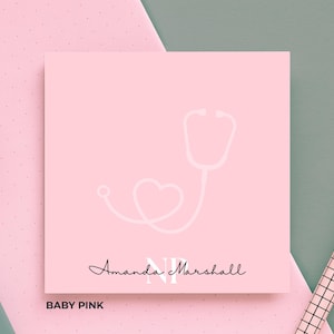 May include: Baby pink notepad with a white stethoscope design, a heart, and the name "Amanda Marshall" in script. The letters "NP" are in a larger font. The words "Baby Pink" are at the bottom.