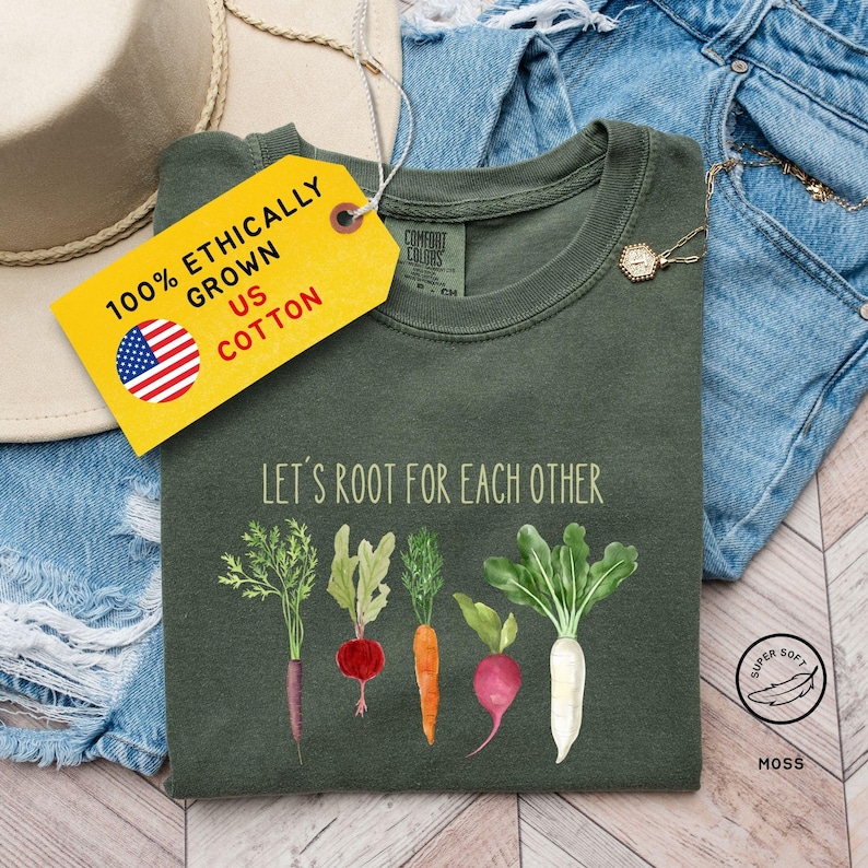 A T-shirt featuring the phrase Let's Root For Each Other in green letters above illustrations of assorted vegetables, including carrots, radishes, and beets. The design aims to celebrate gardening and unity among garden enthusiasts. The shirt is marketed as suitable for gardeners and anyone who loves vintage garden-themed apparel.