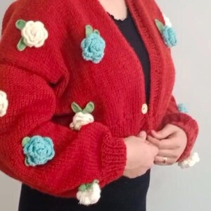 Puede incluir: Un cárdigan de punto rojo con adornos de flores de ganchillo en blanco, azul y verde. El cárdigan tiene cuello en V y cierre de botón.