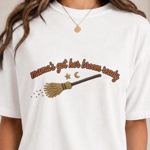 May include: A white short-sleeve t-shirt featuring the brown text "mama's got her broom ready" arched above a graphic of a straw broom, a small star, and a crescent moon. The design is centered on the chest of the crew neck garment.
