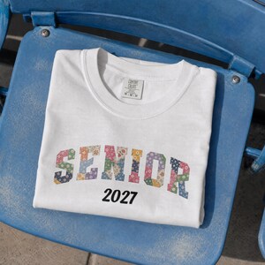 May include: A white short-sleeve t-shirt with a crew neckline. The word SENIOR is printed in a block font with a multi-colored floral pattern. Below the word, the number 2027 is printed in a simple black font. A Comfort Colors brand tag is visible.