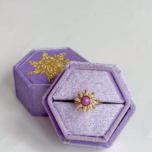 May include: A gold-colored ring with a sunburst design, featuring a central pink gemstone and surrounding clear stones. The ring rests in an open, hexagonal, lavender velvet ring box. A closed box with a gold glitter sun design is visible.