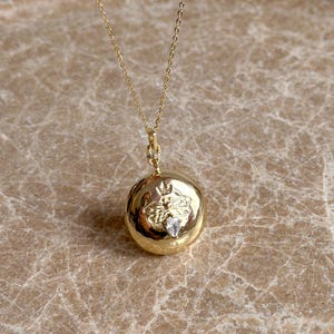 May include: A gold chain necklace with a round pendant featuring a bee design and a small clear crystal. The pendant is on a brown marble surface.