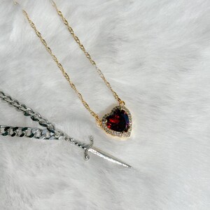 May include: A gold-toned necklace with a heart-shaped pendant featuring a large, red gemstone surrounded by small, clear stones. A silver chain necklace and a small silver dagger are also present.