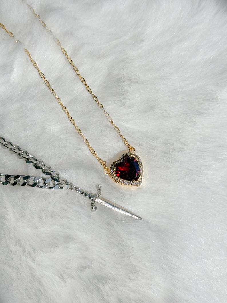 May include: A gold-toned necklace with a heart-shaped pendant featuring a large, red gemstone surrounded by small, clear stones. A silver chain necklace and a small silver dagger are also present.