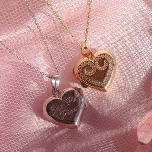 May include: Two heart-shaped pendant necklaces. One is silver with the inscription "Sugar Plum Princess." The other is gold with a decorative design. Both are on delicate chains, set against a pink, textured background.