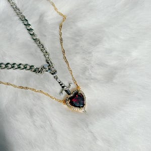 May include: A two-strand necklace featuring a silver chain and a gold chain. The gold chain holds a pendant with a heart-shaped, red gemstone surrounded by small clear stones, with a silver dagger piercing the heart.