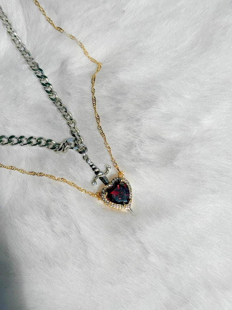 May include: A two-strand necklace featuring a silver chain and a gold chain. The gold chain holds a pendant with a heart-shaped, red gemstone surrounded by small clear stones, with a silver dagger piercing the heart.