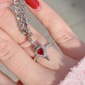 May include: A silver chain necklace with a pendant featuring a red heart with a silver dagger through it. The pendant is attached to a silver ring with a red heart and silver dagger design.