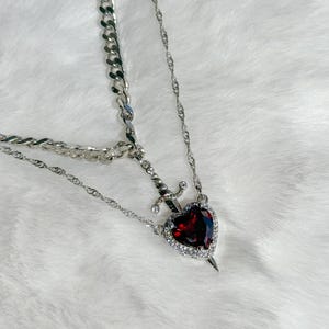 May include: A silver-toned necklace featuring a layered design. The primary pendant is a heart-shaped, deep red gemstone, encircled by clear crystals, with a small sword piercing through it. The necklace has a chain and a curb chain.
