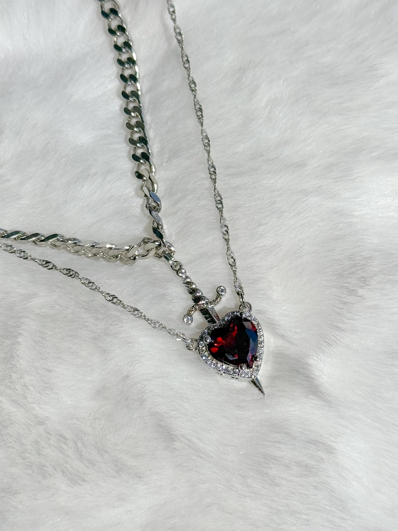 May include: A silver-toned necklace featuring a layered design. The primary pendant is a heart-shaped, deep red gemstone, encircled by clear crystals, with a small sword piercing through it. The necklace has a chain and a curb chain.