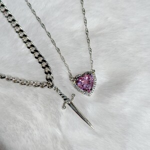 May include: Two silver necklaces. One features a heart-shaped pink gemstone pendant surrounded by small clear stones. The other has a dagger pendant. Both necklaces are on a white, fluffy surface.