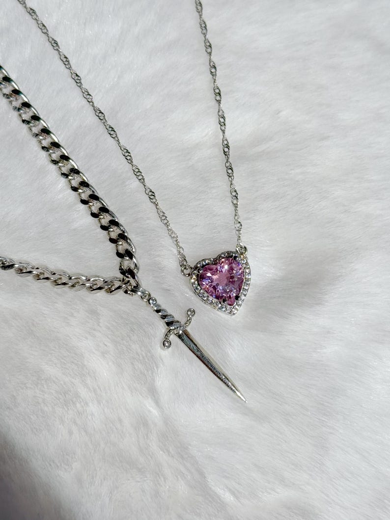 May include: Two silver necklaces. One features a heart-shaped pink gemstone pendant surrounded by small clear stones. The other has a dagger pendant. Both necklaces are on a white, fluffy surface.
