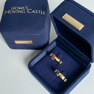 Puede incluir: Dos anillos de oro con piedras preciosas azules y rojas en una caja de terciopelo azul. La caja está etiquetada con "Howl's Moving Castle".
