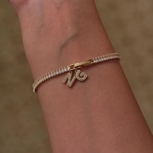 May include: A gold bracelet with a row of clear, rectangular stones. The bracelet has a charm with the letters "NG" encrusted with smaller stones. The bracelet is fastened with a gold clasp.