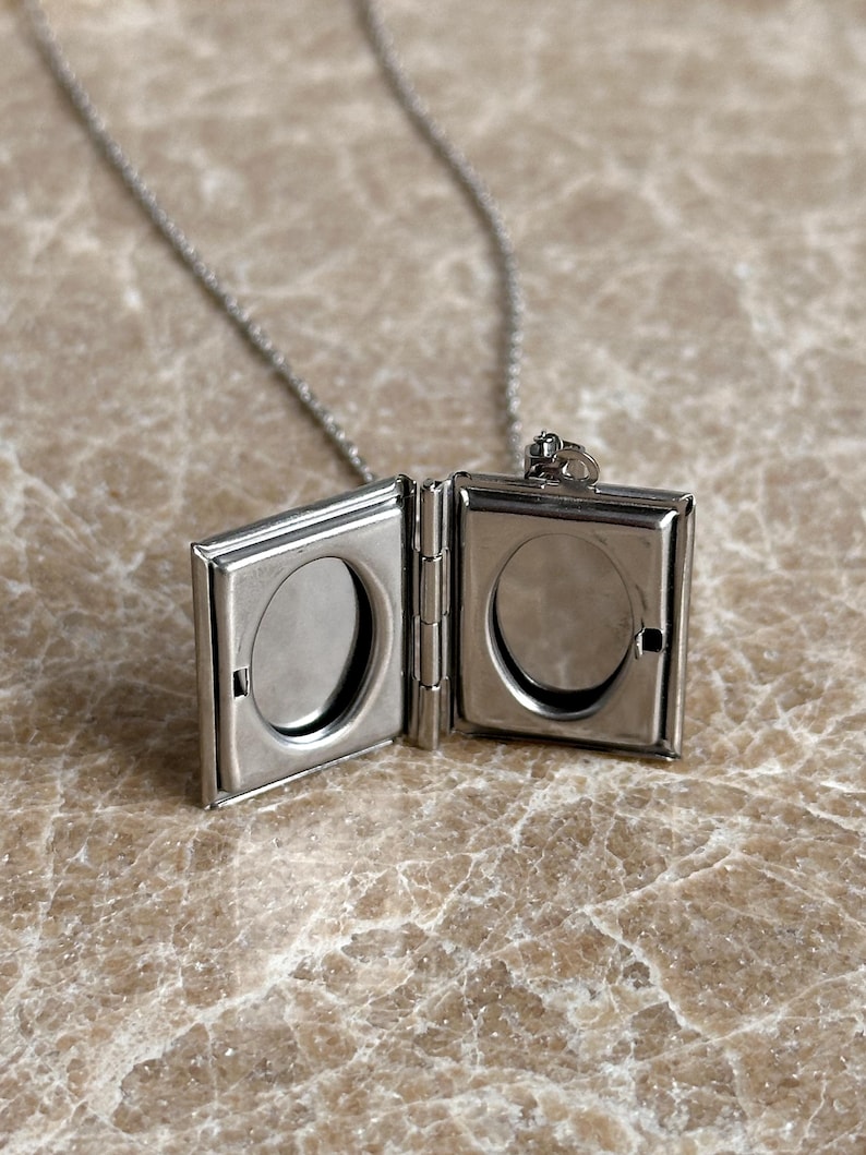May include: A silver chain necklace with a silver locket pendant. The locket is open and has two oval-shaped photo slots.