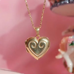 May include: A gold-colored heart-shaped locket pendant on a delicate chain. The locket features an embossed swirl design on the front. The necklace is displayed against a soft pink background, suggesting a romantic or feminine theme.