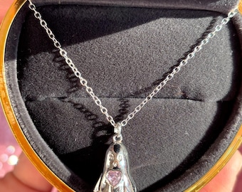 Può includere: Una collana d'argento con un ciondolo a forma di fantasma. Il ciondolo a forma di fantasma ha una gemma a forma di cuore rosa e dettagli neri. La collana è esposta in una scatola a forma di cuore con interno nero.