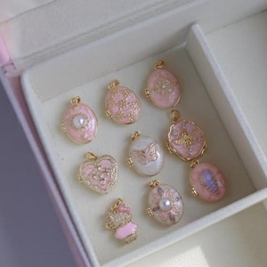 Puede incluir: Una colección de colgantes rosas y dorados de varias formas, incluyendo óvalos, corazones y una magdalena. Algunos colgantes presentan perlas, mariposas y diseños florales. Los colgantes se exhiben en una caja de joyería blanca.