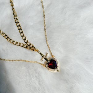 May include: Gold-toned necklace featuring a heart-shaped pendant with a red gemstone center, encircled by clear crystals. A small sword design pierces the heart. The necklace has two chain styles: a curb chain and a delicate rope chain.