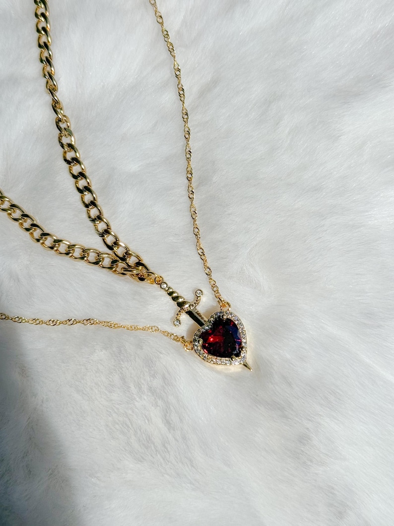 May include: Gold-toned necklace featuring a heart-shaped pendant with a red gemstone center, encircled by clear crystals. A small sword design pierces the heart. The necklace has two chain styles: a curb chain and a delicate rope chain.
