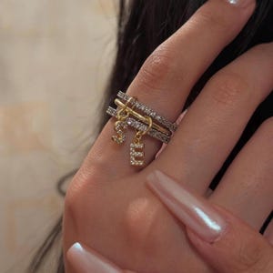 May include: Three stacked rings in gold and silver, embellished with small, sparkling stones. Two gold charms, each with a single letter, "S" and "E", hang from the rings. The rings are designed to be worn together.